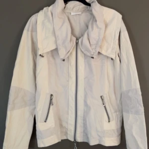 Vit vindjacka från Bonita - Säljer en stilren vit jacka från Bonita med dragkedja och praktiska fickor framtill. Jackan har en hög krage och långa ärmar, perfekt för vår och höst. Den är tillverkad i ett lätt syntetmaterial som gör den bekväm att bära.