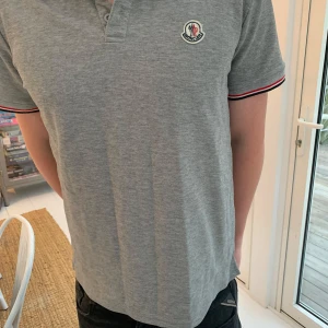 Grå pikétröja från Moncler - Säljer en stilren grå pikétröja från Moncler med korta ärmar och klassisk krage. Tröjan har en broderad logotyp på bröstet och röd-svart randiga detaljer vid ärmslut och krage. Perfekt för en avslappnad men snygg look.