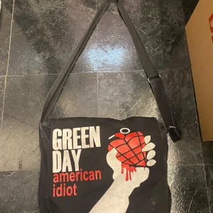 Svart axelväska med Green Day-tryck - Cool svart axelväska med tryck från Green Day's album 'American Idiot'. Väskan har ett stort fack och justerbar axelrem. Perfekt för fans av bandet som vill ha en unik accessoar.