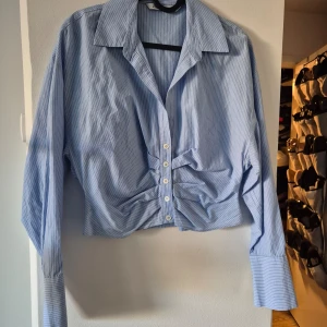 Blå och vit randig skjorta från Zara - Säljer en stilren blå och vit randig skjorta från Zara. Skjortan har långa ärmar och en knäppning framtill med snygg rynkdetalj vid midjan. Perfekt för en chic och avslappnad look.
