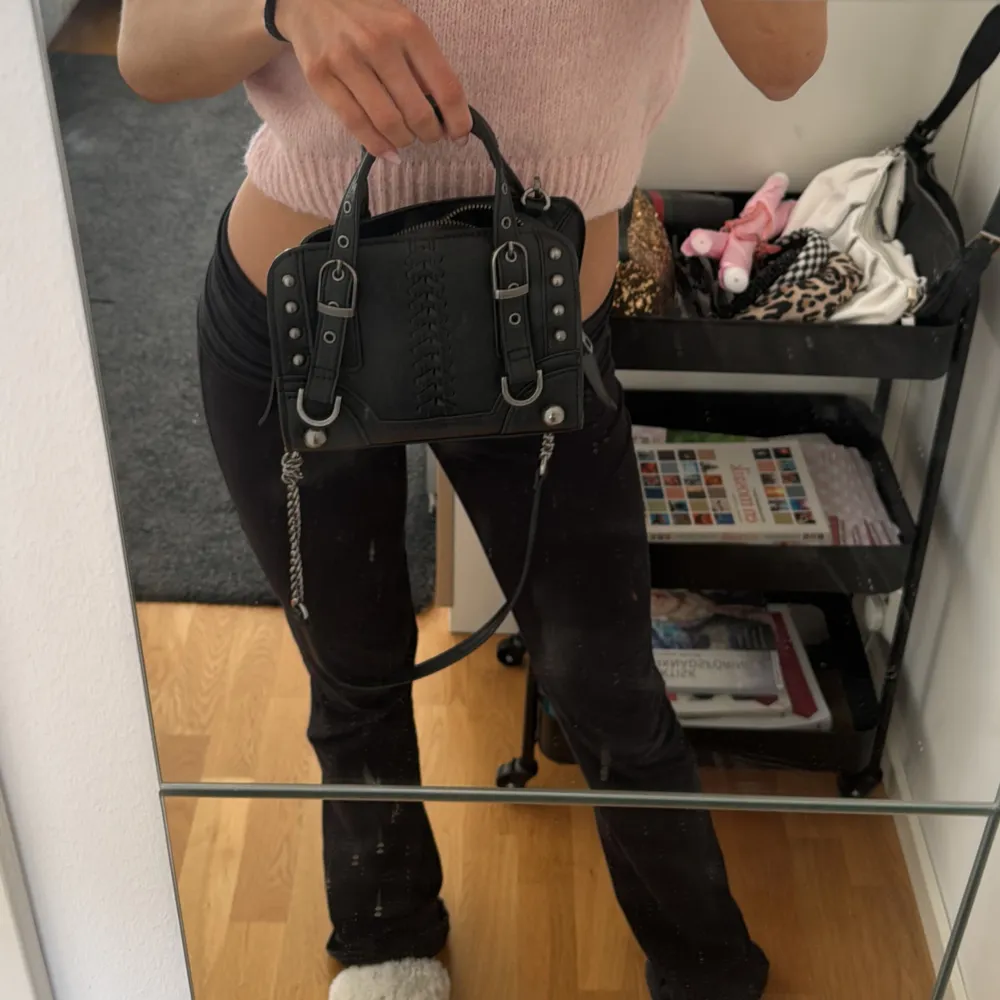 Snygg svart handväska med kedjedetaljer och spännen. Väskan har en dragkedja upptill och dekorativa sömmar på framsidan. Perfekt för att ge din outfit en edgy touch.. Laukut & Käsilaukut.