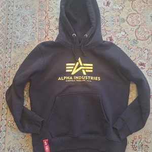 Alpha Industries hoodie - Säljer en svart hoodie från Alpha Industries med guld färgad logga på bröstet. Den har en klassisk känguruficka och justerbar huva med snören. Perfekt för en avslappnad stil.