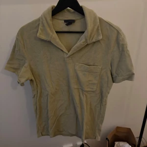 Grön frotétröja med krage - Säljer en stilren grön frotétröja med klassisk krage och korta ärmar. Tröjan har en bröstficka och är perfekt för en avslappnad look. Materialet känns mjukt och bekvämt mot huden.