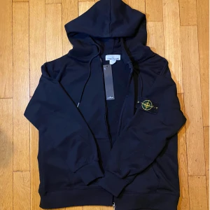 Svart Stone Island hoodie - Säljer en stilren svart hoodie från Stone Island med dragkedja och den ikoniska loggan på ärmen. Perfekt för en casual look. Nytt skick! Säljer pga för liten. Skriv vid fler frågor! 