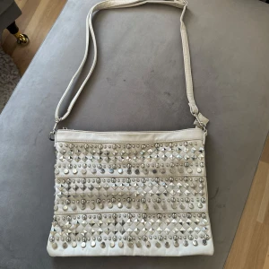 Beige väska - Beige väska med nitar på framsidan och justerbart band. Har ett större innerfack med dragkedja och två mindre öppna innerfack. Använd, så vardagligt slitage, men i bra skick. Har enstaka fläckar i innertyget som syns på bild.