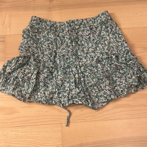 Blommig kjol från Zara - Säljer en söt blommig kjol från Zara i grönt och vitt. Kjolen har en volangdetalj och ett knytband i midjan för en justerbar passform. Perfekt för en somrig look!