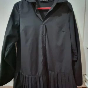 Svart blus från In Vogue - Svart blus från In Vogue med långa ärmar och knappar framtill. Den har en unik plisserad detalj vid nederkanten som ger en extra touch. 