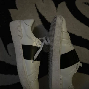 Valentino Garavani sneakers i vitt och svart - Snygga Valentino Garavani sneakers i vitt med en bred svart rem över ovansidan. Skorna har en klassisk design med snörning och en platt sula. Perfekta för en stilren look.