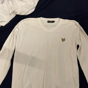 Vit tröja från Lyle & Scott - Säljer en stilren vit tröja från Lyle & Scott med v-ringning och deras ikoniska logga på bröstet. Perfekt för en avslappnad men ändå snygg look. Tröjan har långa ärmar och är gjord i ett mjukt material.