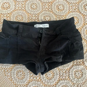 Svarta shorts från Bershka - Snygga svarta shorts från Bershka med låg midja och två knappar framtill. Modellen är tight och har fickor på sidorna. Perfekta för varma dagar när du vill ha en cool och enkel look.