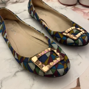 Färgglada ballerinaskor med guldspänne - Unika ballerinaskor med färgstarkt geometriskt mönster i blått, gult, grönt, vinrött och vitt. Snyggt guldigt spänne framtill ger extra edge. Platt sula och rund tå gör dem bekväma och lätta att bära. Perfekta för dig som vill sticka ut med din stil.