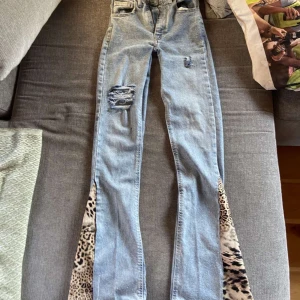 Ljusblå jeans med leoparddetaljer - Unika ljusblå jeans med slitningar och hål på låret samt leopardmönstrade kilar längst ner på benen. Modellen har normal passform och utsvängda ben med coola detaljer som ger en edgy vibe. Perfekta för dig som vill sticka ut lite extra.