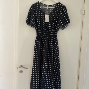 Svart prickig långklänning från Defox - Supersöt svart långklänning från Defox med vita prickar, v-ringning och korta, korta ärmar. Smockad midja ger en snygg siluett och klänningen har ett flowigt fall. Perfekt för dig som gillar retrovibbar och vill sticka ut med ett klassiskt mönster. Ny med prislappen kvar.