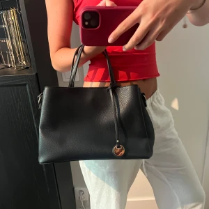 Svart handväska i skinn med gulddetalj - Snygg svart handväska i skinn med stilren design och två handtag. Väskan har en praktisk dragkedja upptill och en rund guldfärgad detalj framtill. 