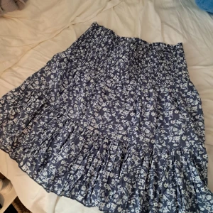 Blommig volangkjol från 157, blå/vit - Supersöt blå kjol med vita blommor från 157. Kjolen har smock i midjan och flera volanglager som ger en flowy känsla. Perfekt för dig som gillar romantisk och chill stil. Lätt och luftig, passar grymt till ett linne och sneakers eller sandaler.