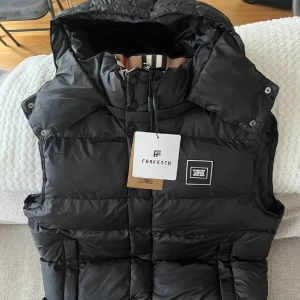 Svart dunväst från Burberry - Snygg svart dunväst från Burberry med hög krage och avtagbar huva. Västen har quiltad design, tryckknappar framtill och en diskret Burberry-logga på bröstet. Perfekt för lager-på-lager och streetstyle.
