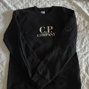 Svart sweatshirt från C.P. Company - Svart sweatshirt från C.P. Company med klassisk logga framtill och den ikoniska linsen på ärmen. Tröjan har rund halsringning och långa ärmar. Perfekt för dig som gillar streetwear och vill ha en stilren look.