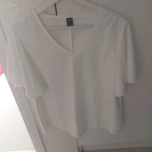 Vit blus med v-ringning från SHEIN - Vit blus från SHEIN med v-ringad hals och korta, luftiga fjärilsärmar. Tillverkad i mjuk polyester med lite stretch för extra komfort. Enkel och stilren design som passar perfekt till jeans eller kjol.