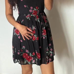 Svart blommig kortklänning - Supersöt svart kortklänning med röda och rosa blommor, markerad midja och lätt utställd kjol. Ärmlös modell med rund halsringning och flowigt tyg som ger en luftig känsla. Perfekt för dig som gillar romantiska vibes och vill sticka ut med färgglada detaljer.
