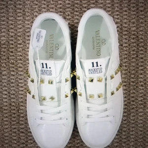 Valentino Rockstud vita sneakers - Snygga vita Valentino Rockstud sneakers med guldiga nitar och klassisk snörning. Skorna har en stilren design med rund tå och platt sula, tillverkade i skinn. Perfekta för dig som vill sticka ut med exklusiva detaljer. Passar 38-39 och är aldrig använda fick dom som present men va inte riktigt min stil Vill gärna bli av med dom så fort som möjligt.❤️