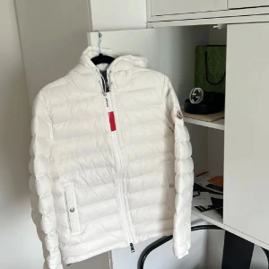 Vit Moncler dunjacka med huva ÄKTA  - Säljer en snygg vit dunjacka från Moncler med huva och dragkedja framtill. Jackan har Moncler-logga på ärmen, snörning vid huvan och diskreta fickor med knappar. Klassisk pufferstil med horisontella sömmar och en röd detalj vid dragkedjan. Perfekt för kalla dagar.