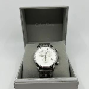 Calvin Klein klocka  - Stilren klocka från Calvin Klein med rund vit urtavla, tre subdials och datumvisning. Armbandet är i silverfärgat meshstål med diskret CK-logga på spännet. Kommer i originalask, perfekt för dig som gillar minimalistisk design och snygga detaljer.