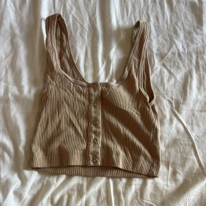 Beige ribbad linnetopp med knappar - Snygg beige ribbad linnetopp med breda axelband och knappar framtill. Croppad modell med djup urringning och stretchigt material som sitter skönt. Perfekt att styla med jeans eller kjol för en chill look.