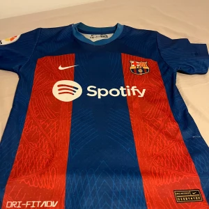 FC Barcelona Lewandowski matchtröja Nike - Säljer en officiell FC Barcelona matchtröja från Nike med Lewandowski och nummer 9 på ryggen. Tröjan är blå och röd med klassiska ränder, klubbmärke, Nike-logga och Spotify som sponsor. Tillverkad i Dri-FIT-material som andas och har korta ärmar.