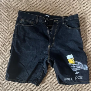 Palace jeansshorts med broderi - Unika svarta jeansshorts från Palace med kontrastsömmar och broderade detaljer. Framsidan har ett skelett som håller ett ölglas och texten 'PAL ACE'. Baksidan har färgglada broderier med peace-märke, blommor och ölglas på bakfickorna. Perfekta för dig som vill sticka ut.
