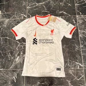 Liverpool Diogo Jota bortatröja Nike S - Liverpool FC bortatröja med Diogo Jota och nummer 20 på ryggen. Tröjan är vit med röda och gula detaljer vid krage och ärmslut, broderad Nike-logga och klubbmärke. Tillverkad i lätt och ventilerande material, perfekt för match eller träning.