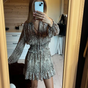 Leopardmönstrad kort klänning från Zara - Superfin kort klänning från Zara med leopardmönster i beige och bruna toner. Klänningen har långärmade puffärmar, smockad midja och volangkant nertill. V-ringad med knytband i halsen och luftig känsla, perfekt för dig som vill sticka ut.