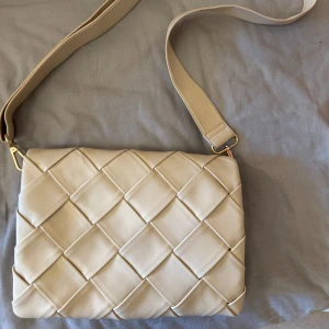 Beige quiltad axelväska i skinnimitation - Säljer en beige axelväska med bred rem och snyggt quiltat mönster. Väskan har flera fack, varav ett med dragkedja, och är gjord i skinnimitation. Perfekt storlek för att få plats med allt du behöver och passar till många olika outfits.