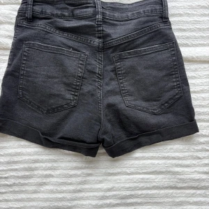 Svarta jeansshorts med hög midja - Svarta jeansshorts med hög midja och klassisk femficksdesign. Shortsen har uppvikta ben.