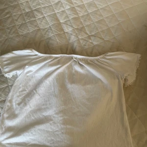 Vit offshoulder blus med spetsdetaljer - Superfin vit offshoulder blus med korta ärmar och spetsdetaljer framtill. Blusen har en söt knytning och små bollar längs ärmsluten. Perfekt för sommarens varma dagar och ger en romantisk vibe till din outfit.