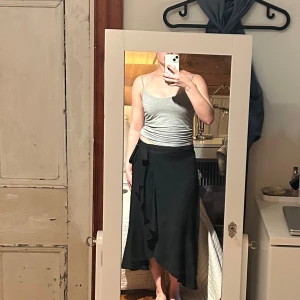 Svart omlottkjol från Gina Tricot - Svart långkjol från Gina Tricot i omlottmodell med knyt i midjan. Kjolen har en lätt volangkant och faller snyggt med en lite asymmetrisk nederkant. Perfekt för dig som gillar en stilren och enkel look.