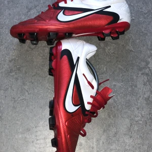 Nike CTR360 röda fotbollsskor - Nike CTR360 fotbollsskor i rött och vitt med svarta detaljer och stora vita swoosh-loggor. Skorna har snörning, platt sula med fasta dobbar och är tillverkade i syntetmaterial. Perfekta för dig som vill sticka ut på planen med stilrena och sportiga skor.