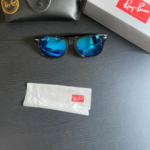 Ray-Ban Wayfarer svarta med blå spegelglas - Snygga Ray-Ban Wayfarer solglasögon med svart båge och blå spegelglas. Klassisk fyrkantig form och ikonisk logga på linsen. Kommer med originalfodral och putsduk. Perfekta för att sticka ut i solen!
