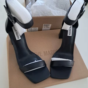 Svarta klackade sandaler Steve Madden - Stilrena svarta sandaler från Steve Madden i skinn, bred rem över foten och justerbart ankelband med rund spänne. Klackarna är blockformade och ger en modern look. Perfekta för dig som gillar minimalistisk och trendig stil.