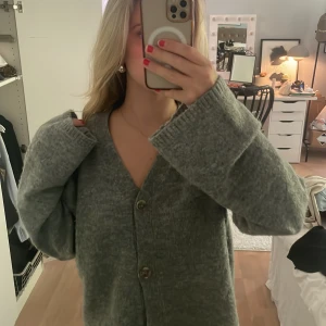 Grå stickad kofta med knappar - Mysig grå stickad kofta med v-ringning och stora knappar framtill. Koftan har långa ärmar och en loose passform som ger en chill vibe. Perfekt att slänga på sig över en t-shirt eller linne när det är lite kyligt.