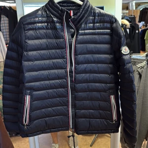 Svart pufferjacka från Moncler - Snygg svart pufferjacka från Moncler med klassisk quiltad design och ståkrage. Jackan har dragkedja framtill, två fickor med dragkedja och Moncler-logga på ärmen. Röd, vit och blå randiga detaljer vid dragkedjorna ger en sportig touch.
