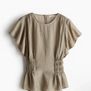 Beige blus med volangärm och smock - Säljer en beige blus med korta, vida volangärmar och smockdetaljer i midjan som ger en snygg form. Blusen har rund halsringning och är tillverkad i ett lätt, krinklat tyg som känns luftigt och skönt. Perfekt för dig som gillar en avslappnad men ändå trendig stil.
