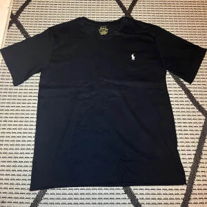 Svart t-shirt Polo Ralph Lauren S - Svart t-shirt från Polo Ralph Lauren i custom slim fit. Klassisk design med korta ärmar och rund hals. Ikoniska broderade loggan i vitt på bröstet. Tillverkad i mjuk bomull som känns skön mot huden. Perfekt för en clean och stilren look. Endast använd 1 gång!