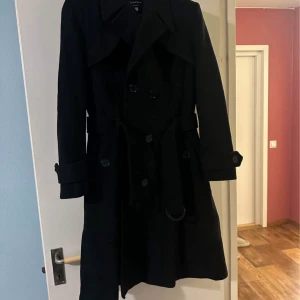 Svart trenchcoat från De la Crème - Svart trenchcoat från De la Crème med dubbelknäppning, bälte i midjan och snygga detaljer vid ärmsluten. Jackan är tillverkad i en mix av ull, polyester och kashmir, vilket gör den både varm och stilren. Perfekt för dig som vill ha en klassisk och tidlös look.