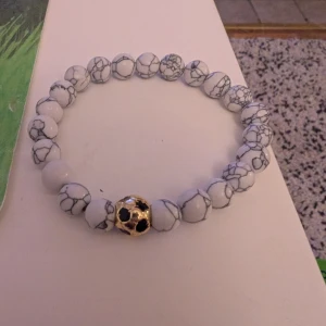 Vitt pärlarmband med guldig fotbollspärla - Stilrent armband med vita marmorerade pärlor och en unik guldig pärla formad som en fotboll. Perfekt accessoar för dig som gillar sportig stil och vill sticka ut lite extra. Elastiskt band gör det enkelt att ta på och av.