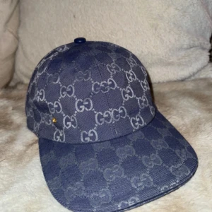 Blå keps från Gucci med GG-mönster - Snygg blå keps från Gucci med ikoniskt GG-mönster över hela kepsen. Tillverkad i bomullstyg med klassisk böjd skärm och diskret guld-detalj på sidan. Insidan är ljus och har tydlig Gucci-label. Perfekt för dig som vill sticka ut med en lyxig accessoar.