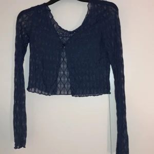 Mörkblå mesh bolerotopp - Säljer en mörkblå, transparent bolerotopp med lång ärm och croppad passform. Toppen har ett diskret mönster i tyget och knäpps framtill. Perfekt att styla över ett linne eller klänning för en trendig look.