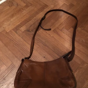 Brun axelväska i skinn - Snygg brun axelväska i skinn med justerbar rem. Väskan har en enkel och stilren design med mjuka former och diskreta sömmar på framsidan. Perfekt storlek för att få plats med det viktigaste och passar till många olika outfits.
