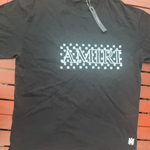 Svart AMIRI t-shirt med tryck - Svart t-shirt från AMIRI med stort vitt logotryck och mönstrade detaljer på bröstet. Klassisk rund halsringning och korta ärmar. Materialet är mjuk bomull och passformen är normal. Perfekt för dig som gillar streetwear med exklusiv känsla.