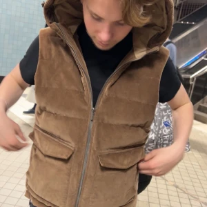 Brun manchesterväst med huva - Säljer en brun dunväst i manchester med stor huva och två fickor framtill. Västen har dragkedja och en lite boxig passform som ger en chill vibe. Perfekt att slänga över en hoodie eller t-shirt för extra stilpoäng.