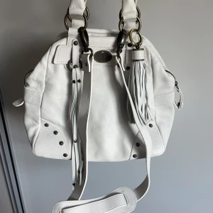 Vit handväska med fransar  - Vit handväska från Maria Montazami i mjukt skinn med coola metalldetaljer och fransar framtill. Väskan har både handtag och axelrem, dragkedja på sidorna och snyggt mönstrat innerfoder med logga. Perfekt för dig som gillar stilrena och coola accessoarer💗💗nypris ca 2000, säljer för 600 eller högstbjudande💗